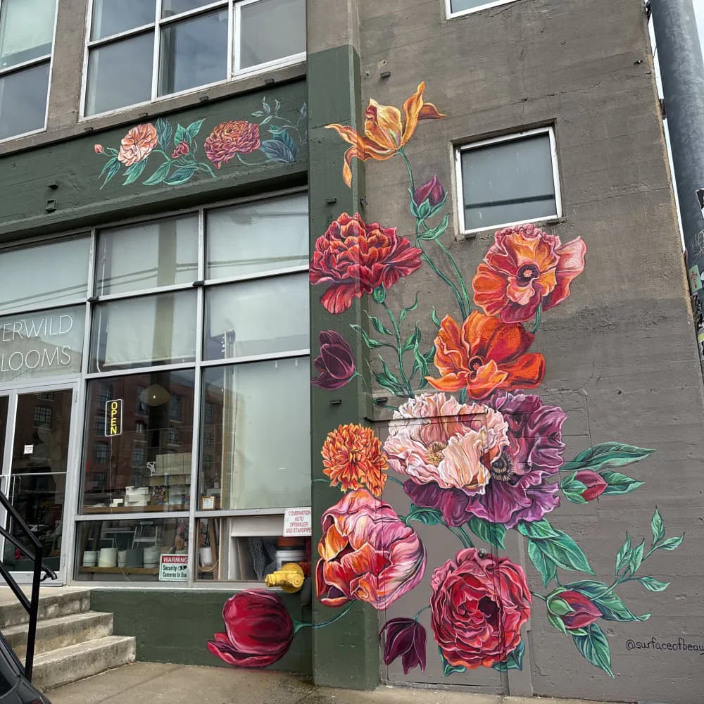Mural by Natasha May Platt featuring cascading botanical illustration of pink, red, orange, and purple flowers with green foliage on dark building facade.