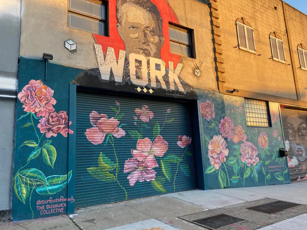 Storefront with blue metal shutter door decorated with pink and purple painted flowers; red and white "WORK" signage and portrait above.