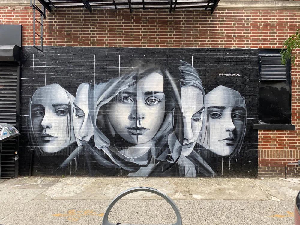 Black and white mural on brick wall featuring three overlapping portrait heads with hooded faces, rendered with fine line-work detail.