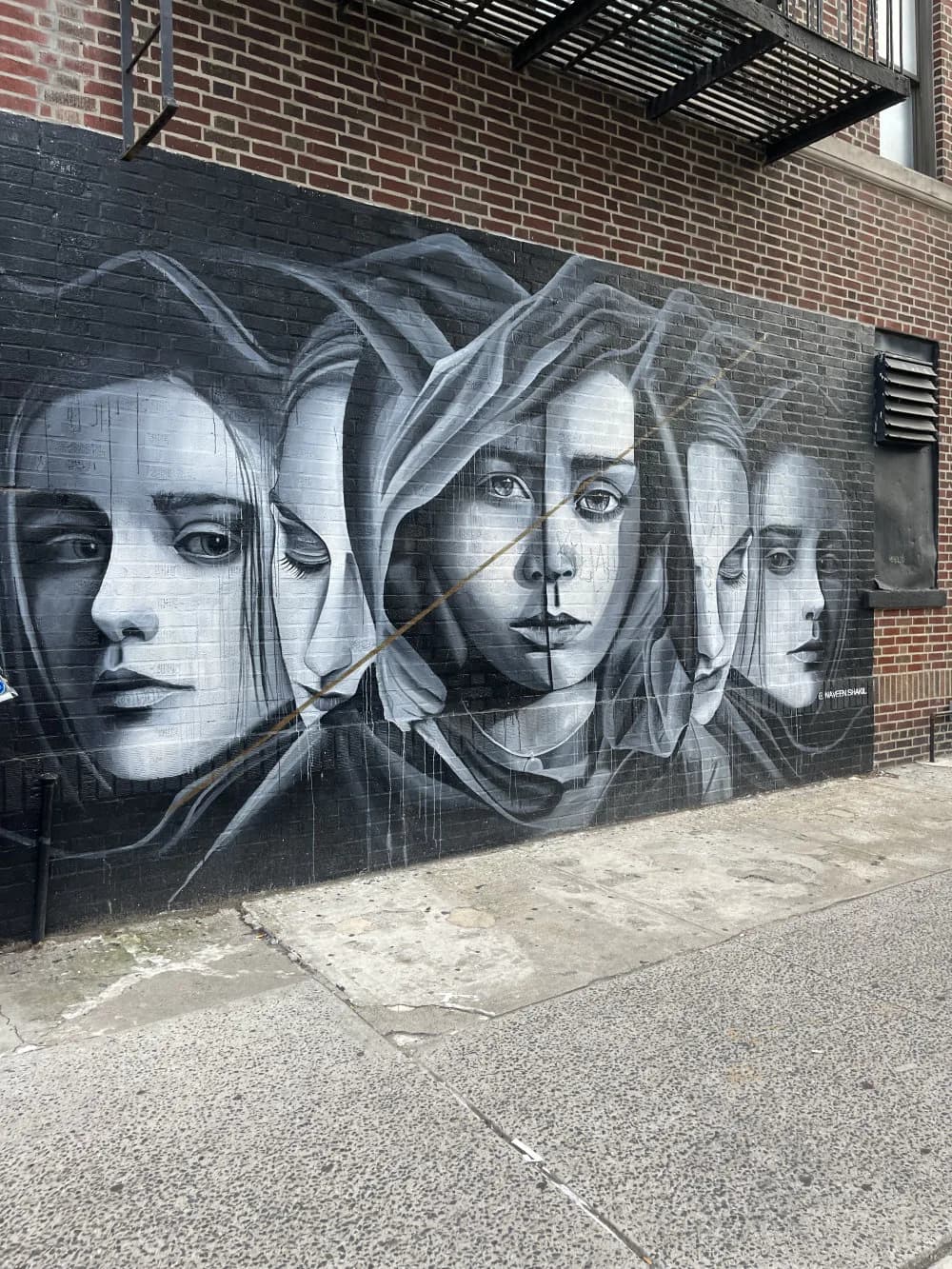 Black and white mural on brick wall depicting three overlapping female faces with draped fabric, executed in detailed grayscale.