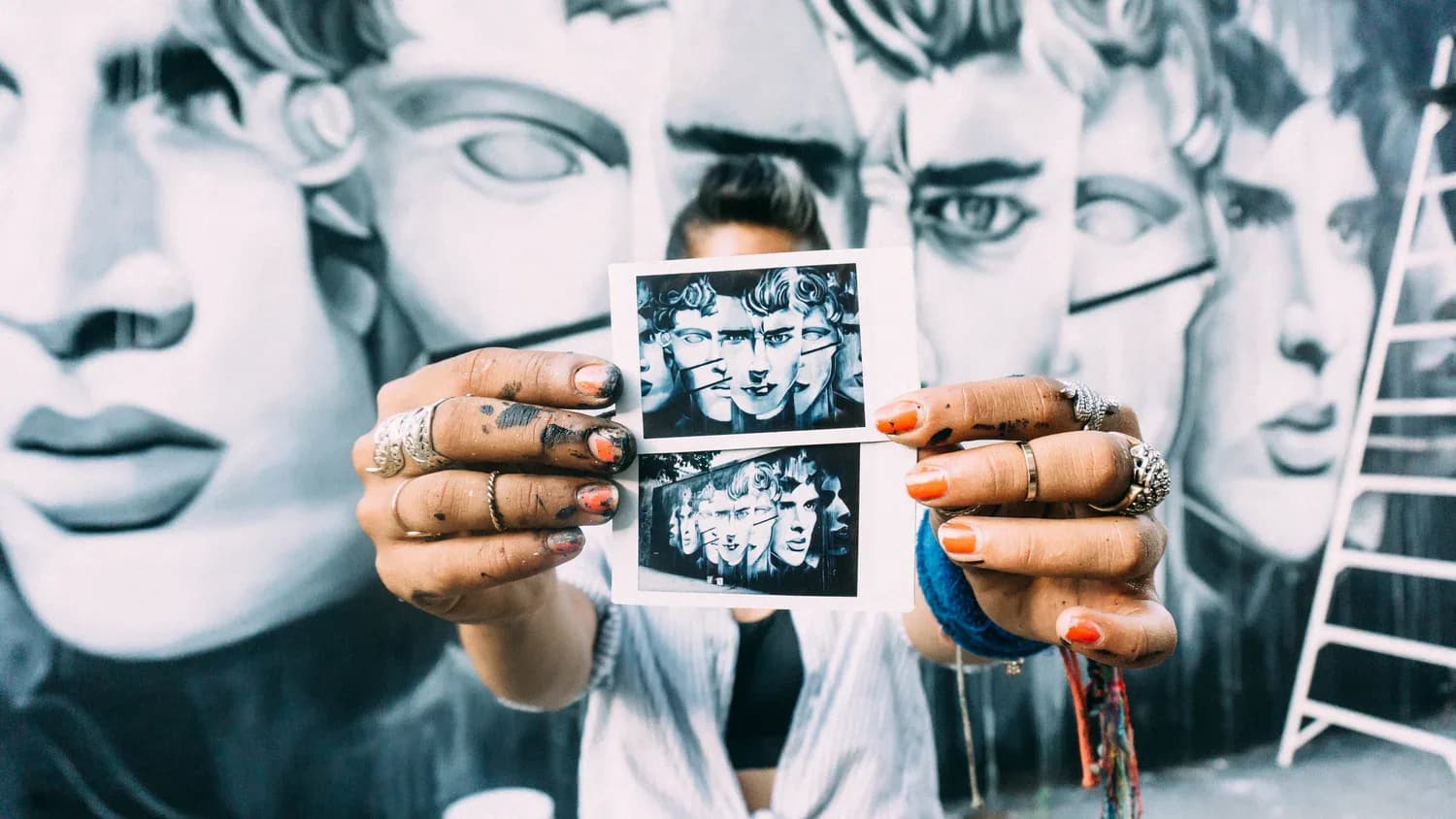 Person's hands holding small black and white Polaroid photographs against a large mural background of multiple portrait faces.