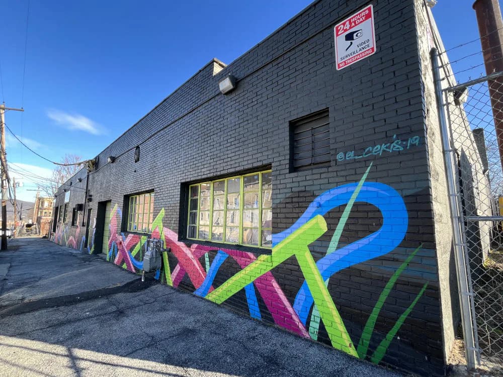 Large-scale mural with bold pink, blue, and green geometric lettering and abstract shapes on brick building storefront.