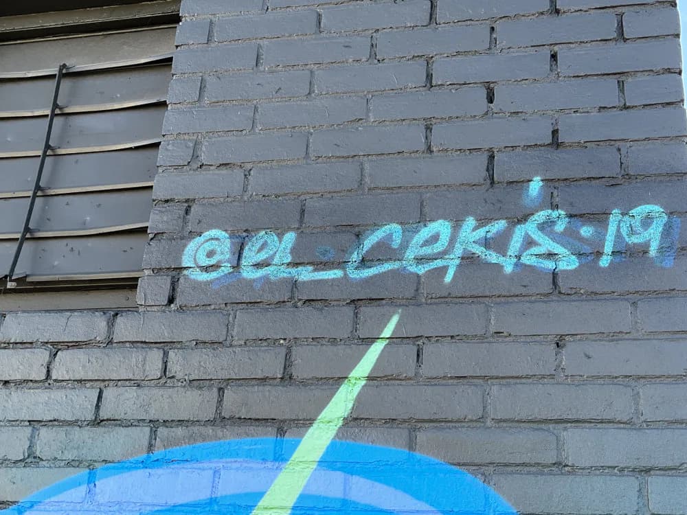 Detail of mural with cyan spray-painted text and blue and green geometric design on gray brick wall.