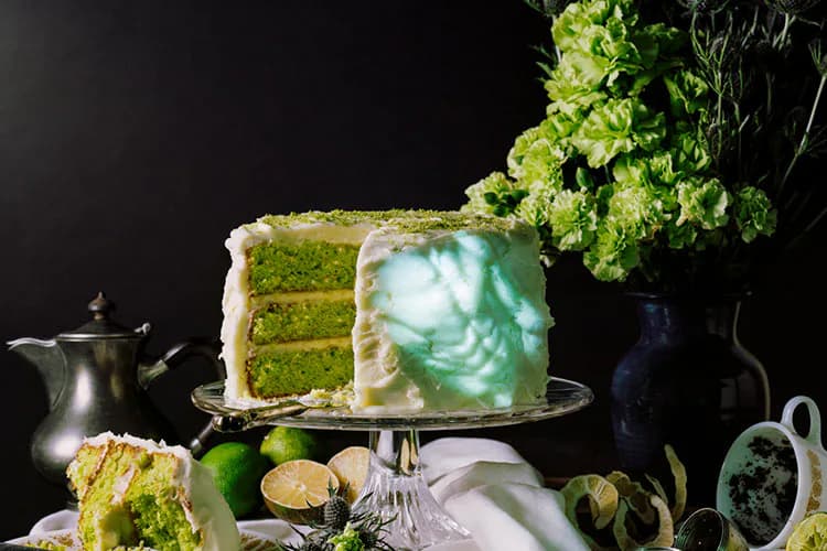 Multi-layered green cake on a pedestal with white frosting, surrounded by fresh limes, herbs, and vintage tableware against a dark background.