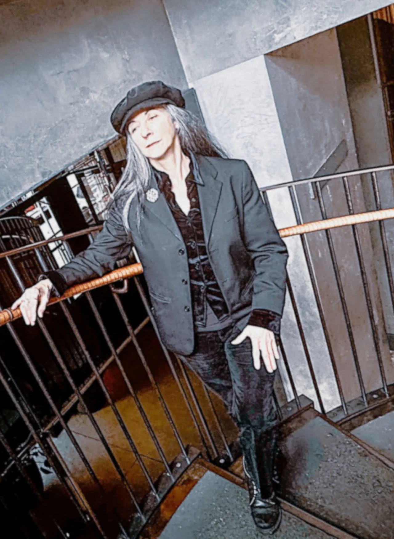 Person wearing a dark blazer and cap leaning against a railing in an industrial stairwell setting, looking at camera.