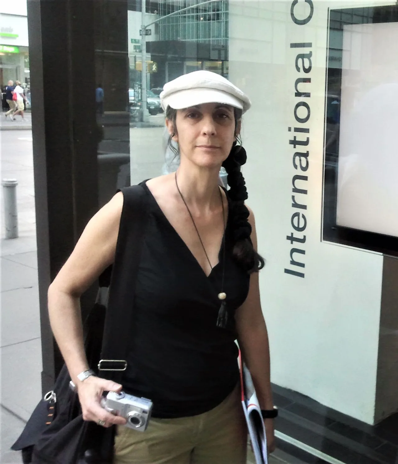 Woman in a white cap and black sleeveless top holding a camera, standing in front of a storefront with 'International' signage.