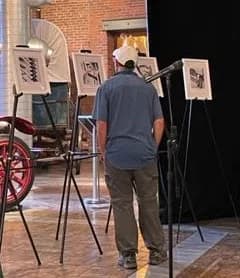 Person viewing artwork displayed on easels in a gallery space with brick wall and bicycle visible in background.