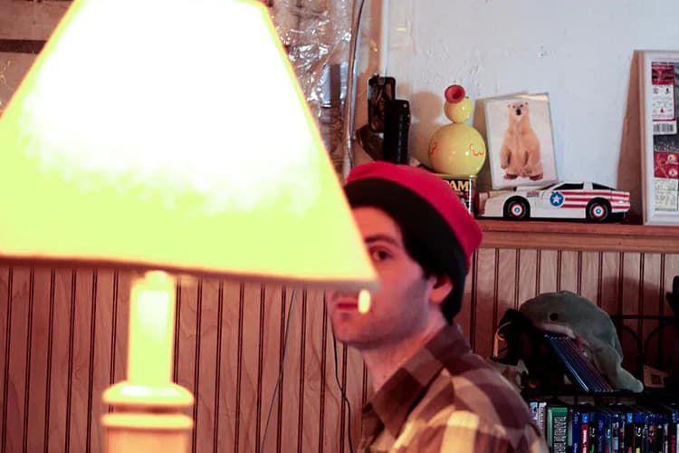Person in profile wearing a red winter hat, illuminated by a bright yellow lamp in an indoor room with shelves and toys.