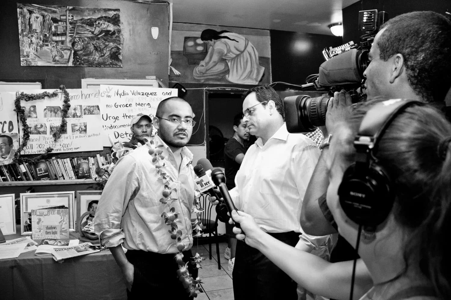 Black and white photograph of a person speaking into a microphone surrounded by journalists and cameras in an indoor space with posters on walls.