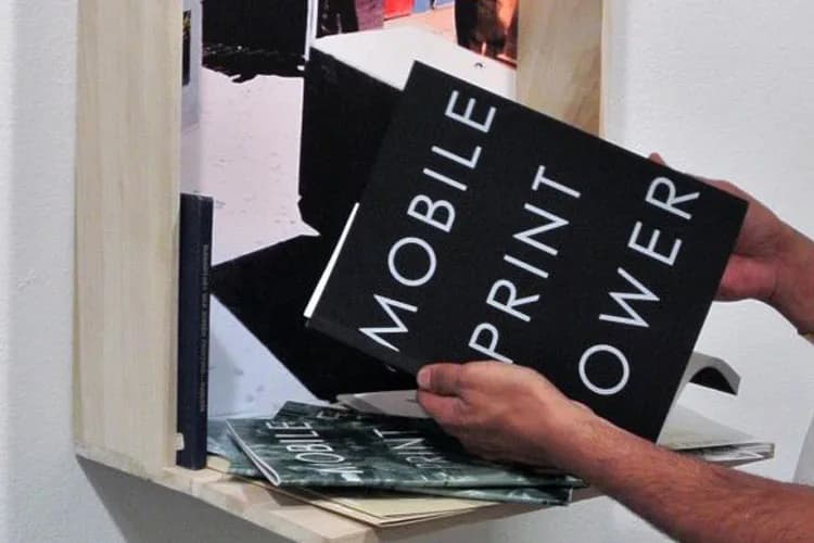 Hands hold a black book cover reading 'Mobile Print Power' beside a wooden shelving installation with printed imagery, part of Patrick Rowe's printmaking work