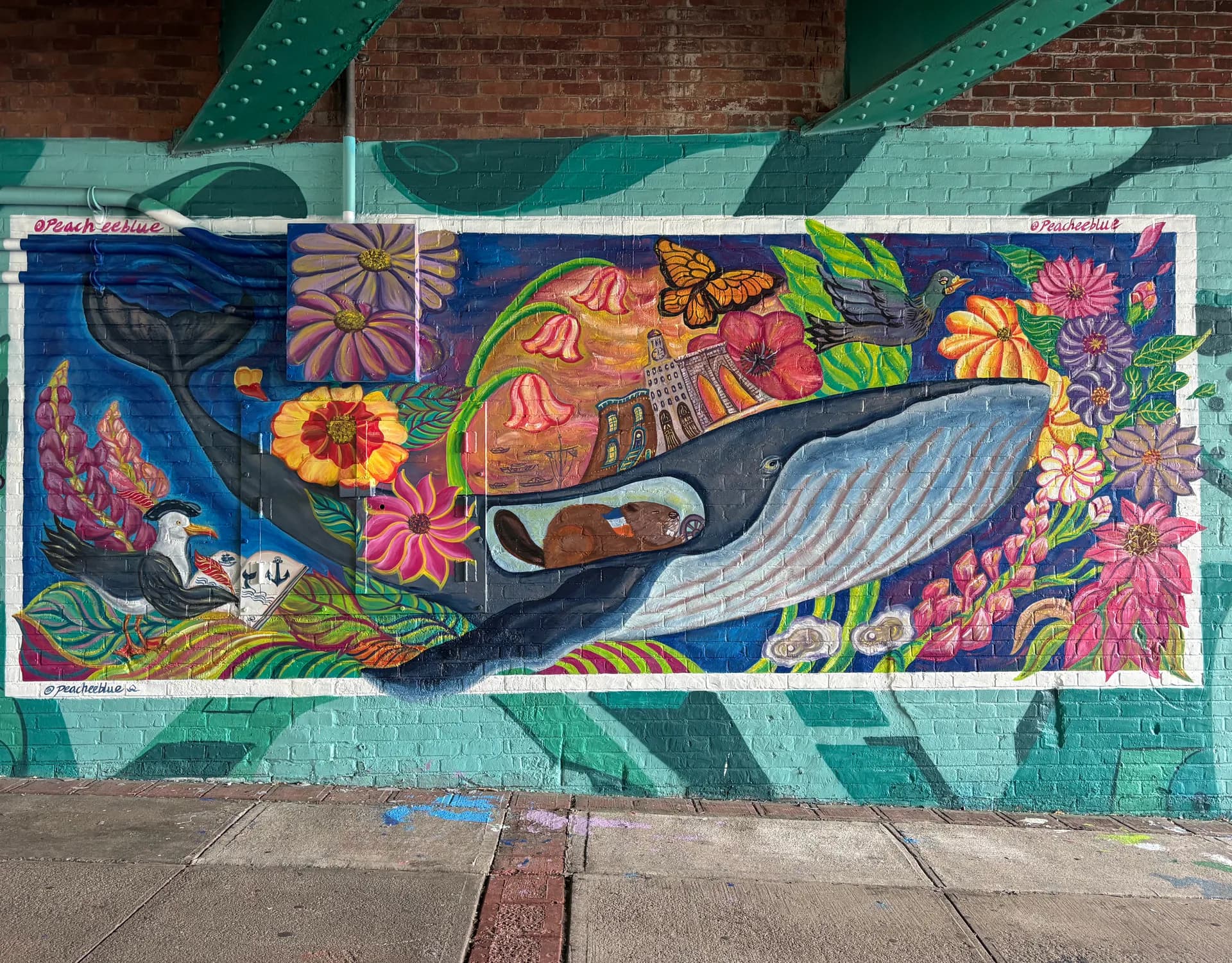 Large-scale street mural featuring a whale surrounded by colorful flowers, a seagull, butterfly, beaver, and Brooklyn landmarks on a teal brick wall