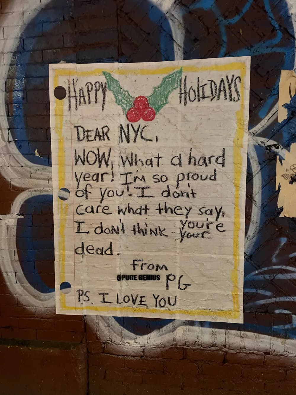 Handwritten holiday card on cream paper with yellow border, addressed to NYC with encouraging message signed by Pure Genius, mounted on brick wall with blue graffiti.