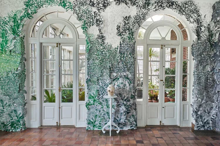 Two white arched doors with glass panels flanked by climbing ivy on a textured wall, installation by Rachel Sydlowski merging architecture with live vegetation.