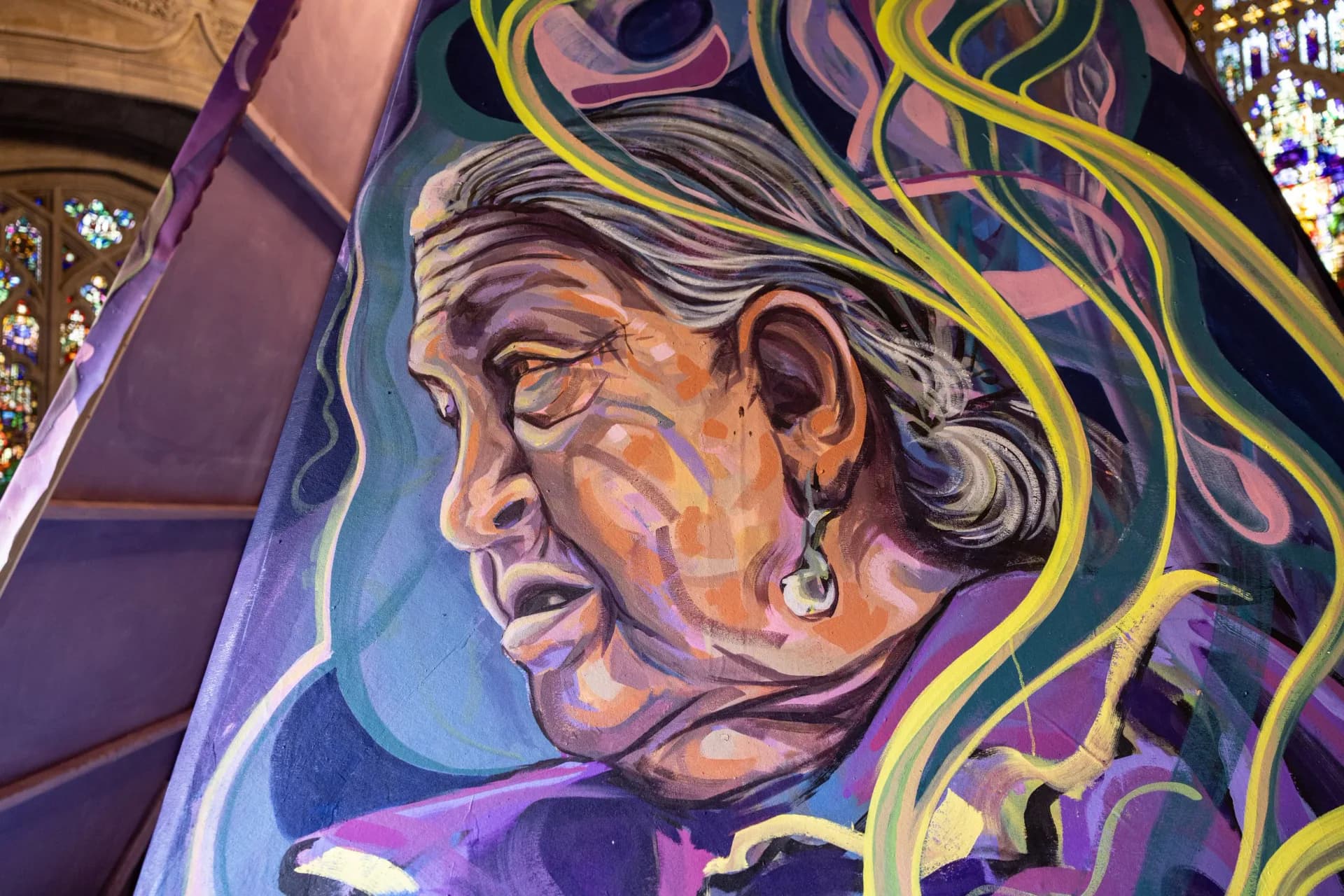 Detail of painted figurative panel showing an elderly woman in profile with flowing yellow and teal hair against purple background, part of cathedral installation