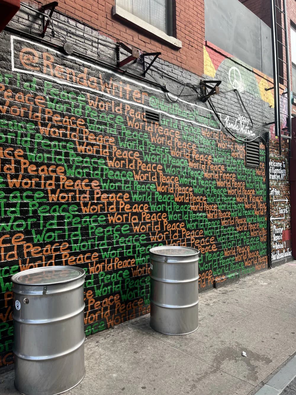 Brick wall mural featuring overlapping text reading "World Peace" in orange, green, and red lettering on contrasting colored sections, with metal bollards in foreground.