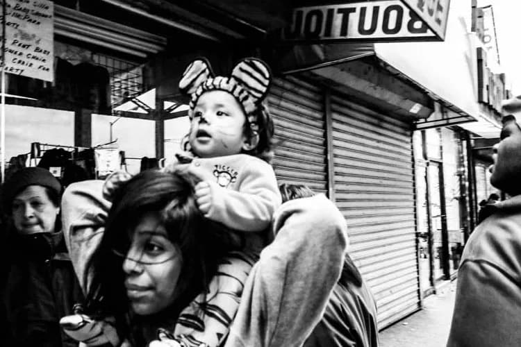 Black and white photograph of a woman holding a young child on a crowded street with storefronts, capturing an intimate moment in an urban setting.
