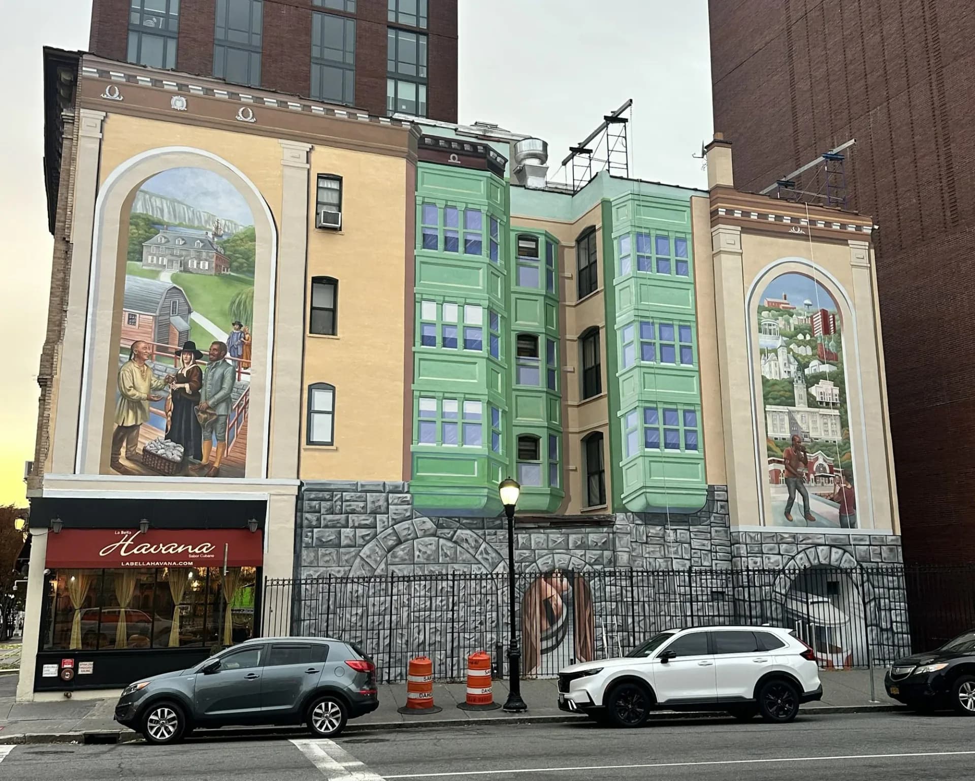 Trompe-l'oeil mural by Richard Haas featuring two large figurative scenes in sepia tones flanking a green-painted building section, with street-level storefront.