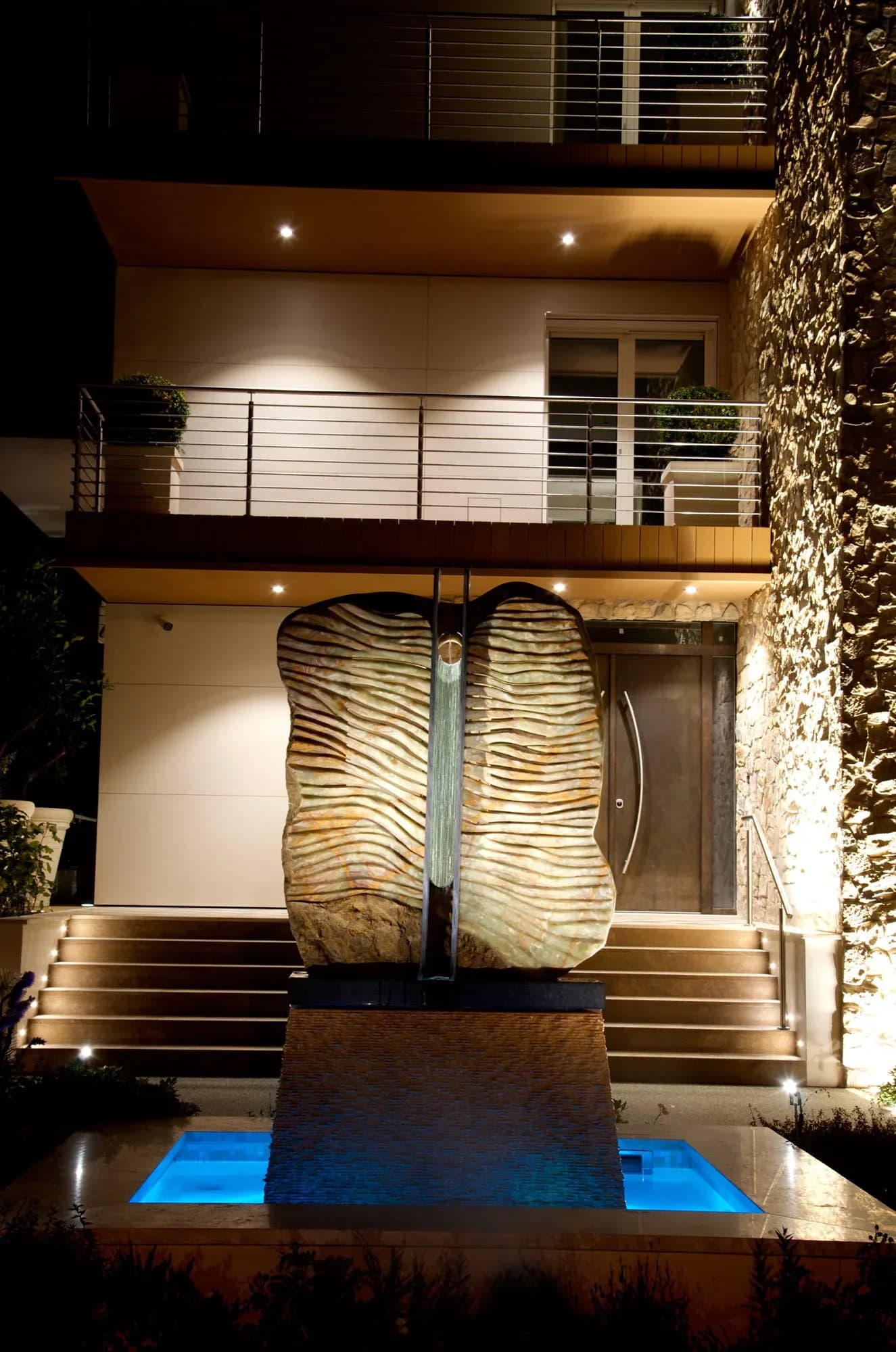 Large onyx sculpture with rippled surface illuminated at night against a residential building with water basin