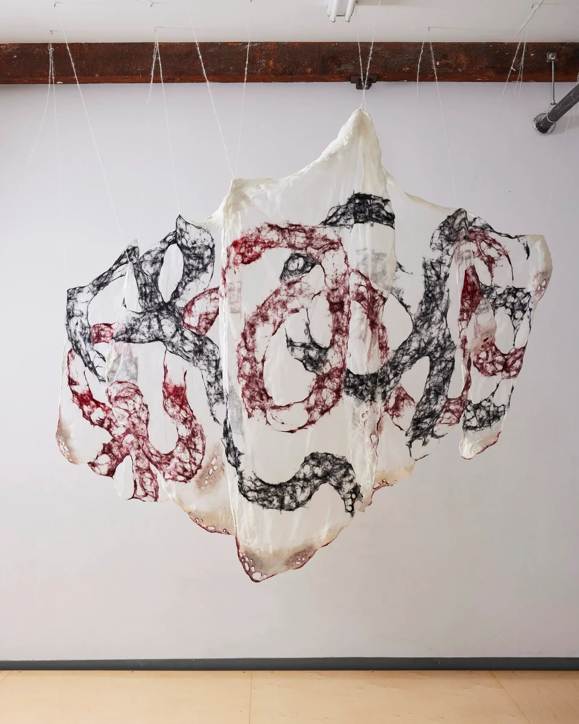 Large-scale hanging felt installation with black and crimson fiber patterns suspended from ceiling beams in gallery space