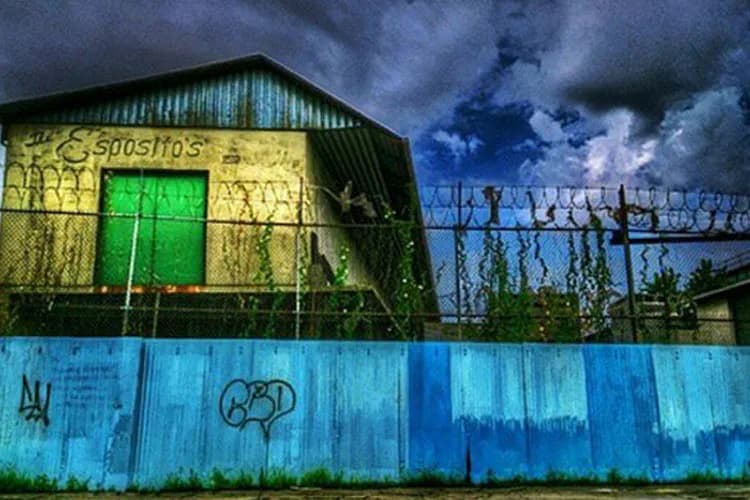 Photograph by Scott Hisley depicting an industrial building with yellow corrugated metal upper section, green door, and blue metal fence barrier below, set against a dramatic cloudy sky.