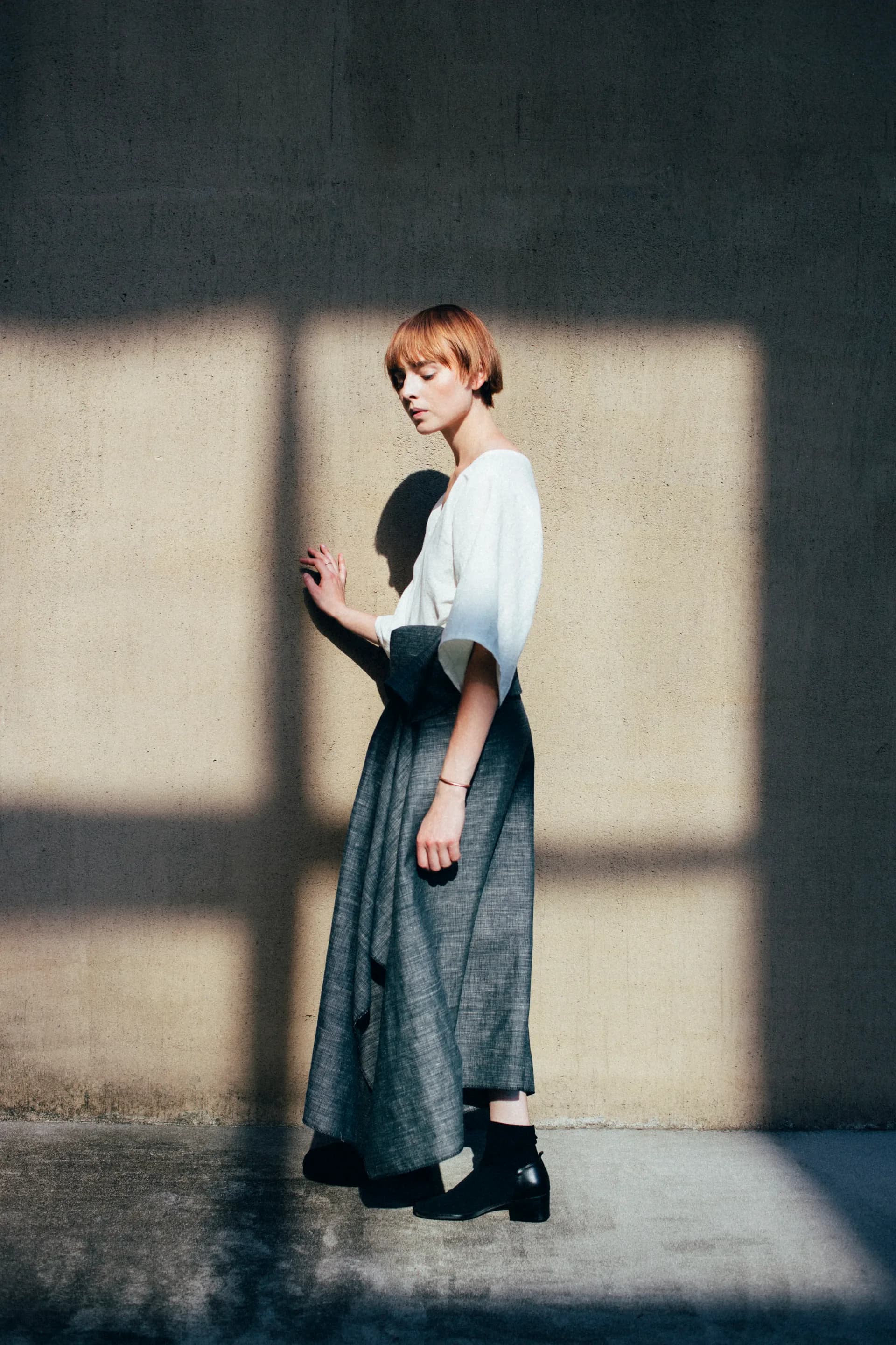 Fashion photography: model in white blouse and grey draped skirt against concrete wall with dramatic window shadow