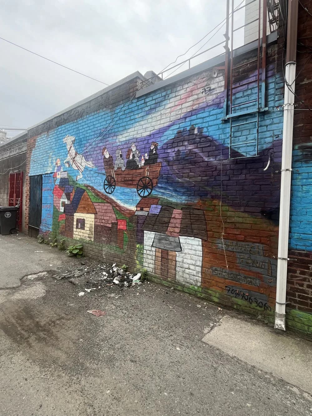 Colorful mural on brick wall depicting figures in a hot air balloon above a patchwork landscape with blue sky, created by Shalom Burkis in Crown Heights.
