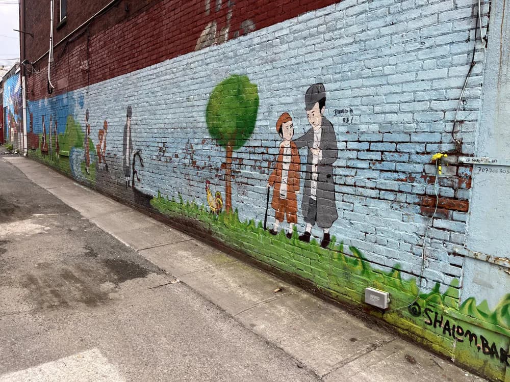 Mural on light blue brick wall showing three figures standing beside a stylized green tree, with grass at the base.