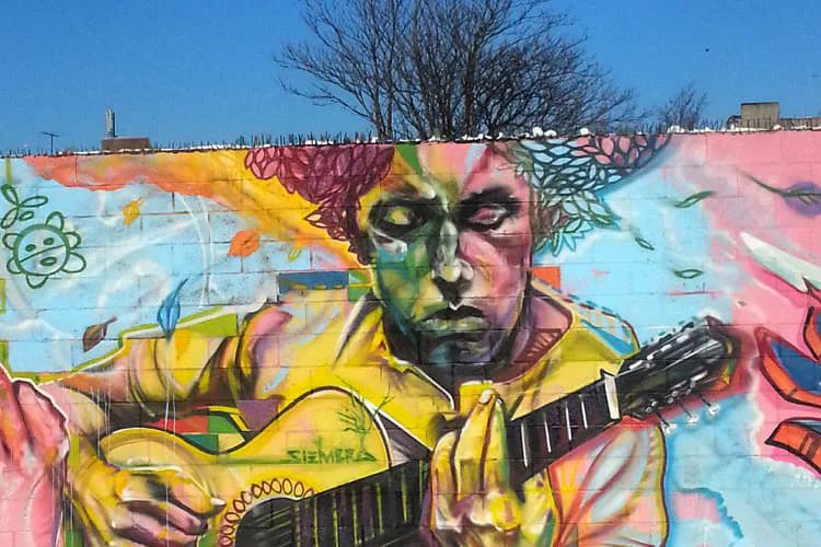 Vibrant mural by SIENIDE featuring a stylized face in yellow, green, and pink tones playing an acoustic guitar against a colorful background with abstract elements.