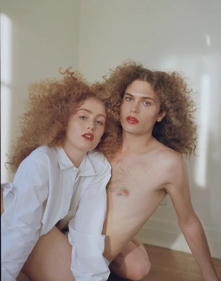 Two figures with matching voluminous curly hair and red lips in intimate pose