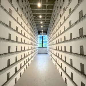 Photograph of a minimalist storage or archive interior with symmetrical rows of shelving units lining both sides and bright natural light from a skylight.