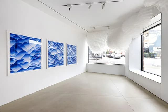 Gallery installation view featuring three blue wave-pattern artworks on white wall and a white sculptural cloud-like installation hanging from ceiling