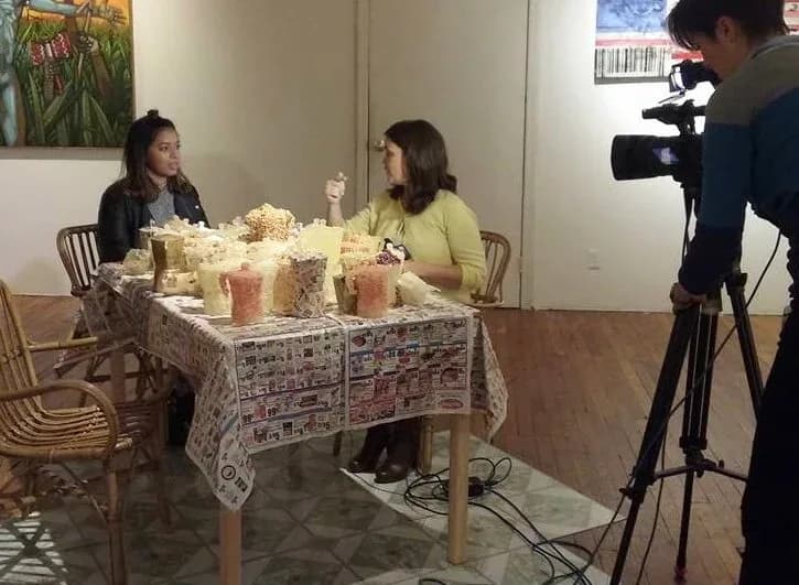 Two people seated at table with patterned tablecloth and popcorn containers during video filming or interview; cameraman visible on right in indoor gallery space.