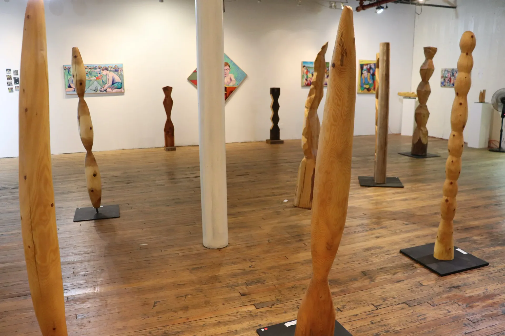 Installation view of multiple tall carved wood sculptures in a gallery space with paintings on walls