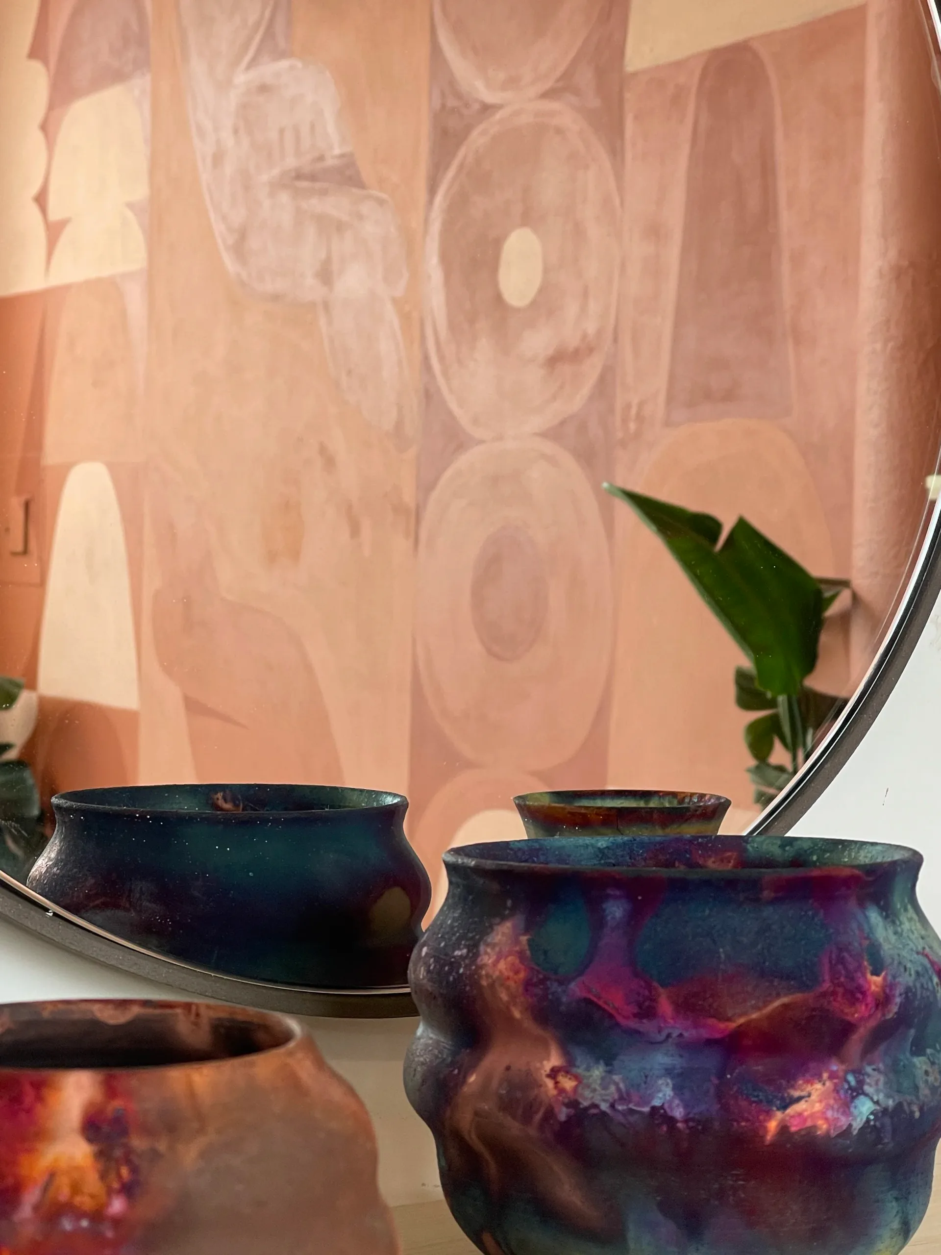 Raku-fired ceramic vessels with iridescent purple, teal, and copper glazing displayed in front of an abstract mural
