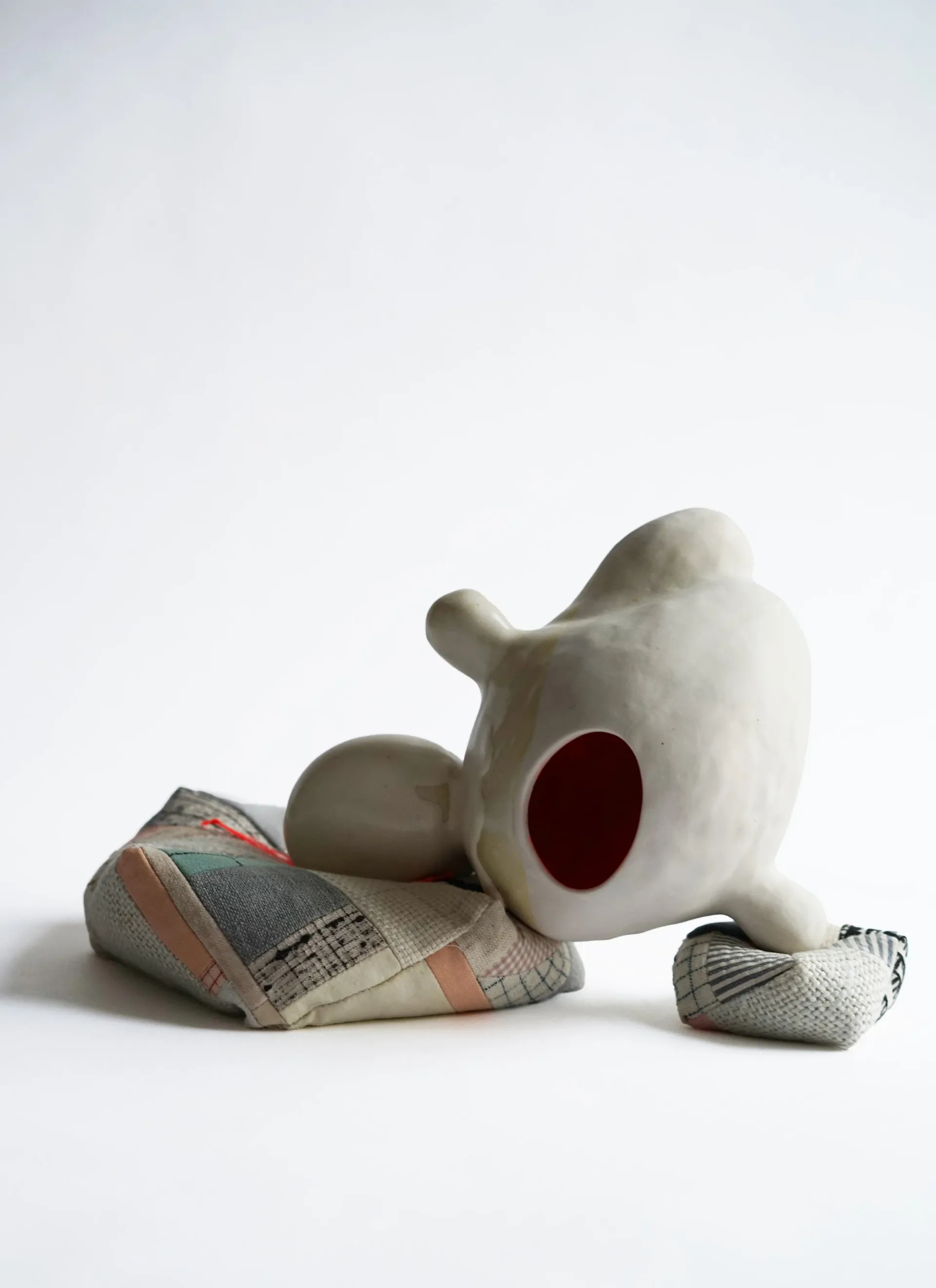 White ceramic sculptural form with deep burgundy circular opening, seated on patterned fabric remnants in gray and cream tones.