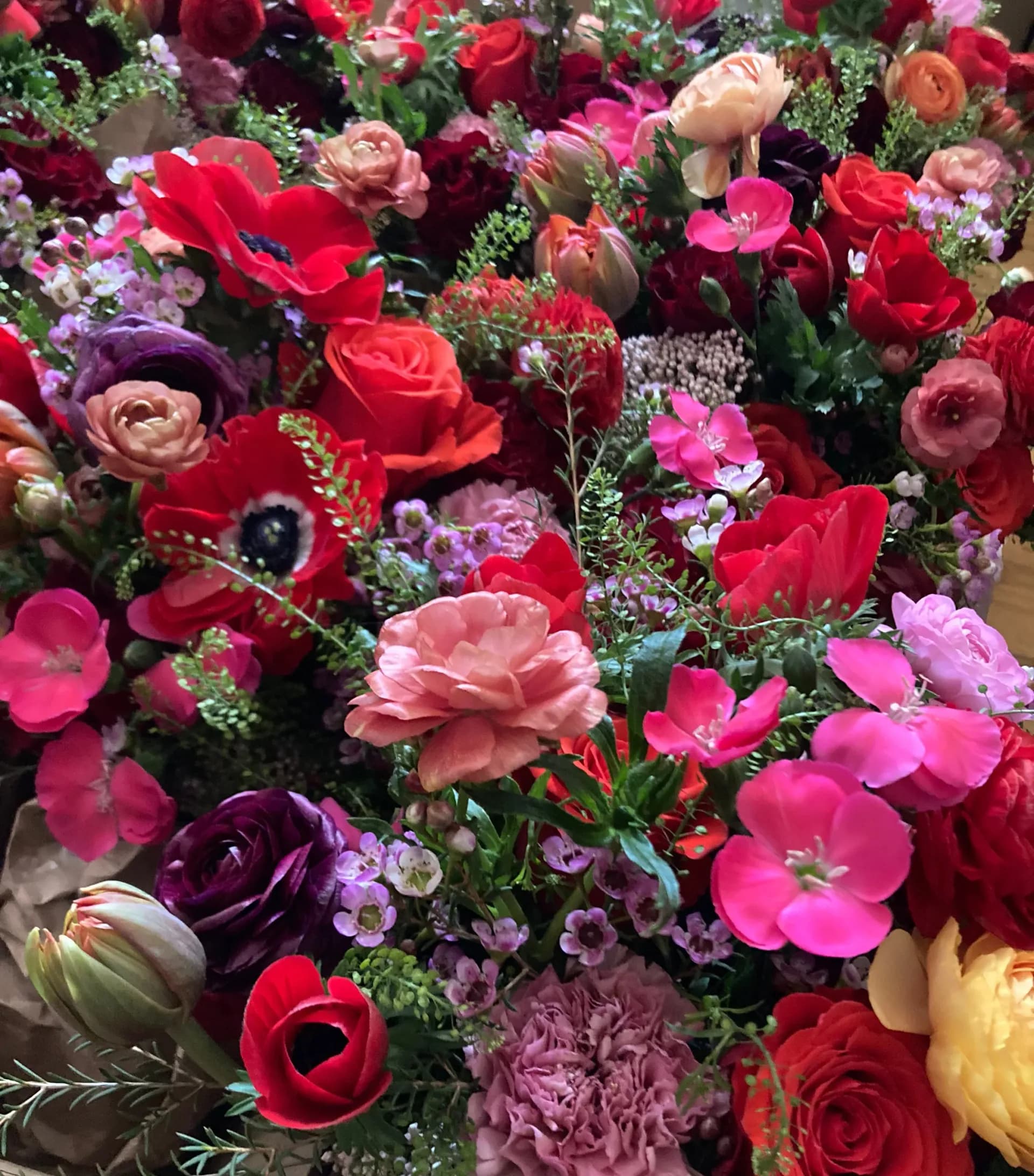 Dense, colorful floral arrangement featuring red, pink, magenta, and deep purple roses, carnations, and spray flowers with white baby's breath and green foliage.