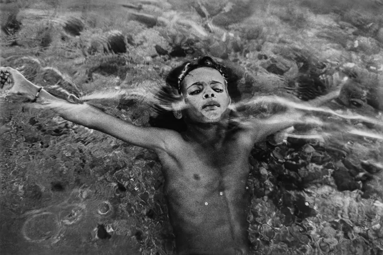 Black and white photograph of a shirtless young child lying in shallow water with arms spread, gazing upward at camera.