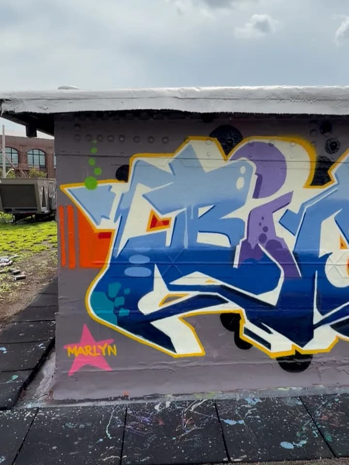 Graffiti mural on gray wall featuring blue, purple, and yellow wildstyle letters with 3D effect and 'MARILYN' signature in pink.