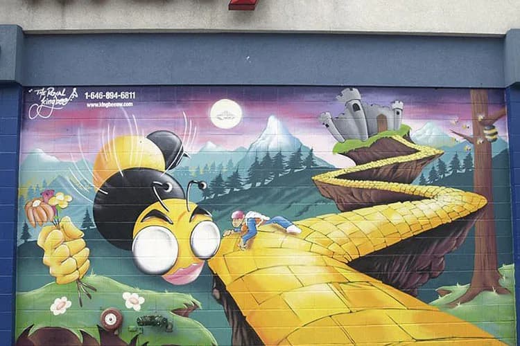 Vibrant mural by The Royal 'Kingbee' featuring a giant yellow-and-black bee with large expressive eyes overlooking a surreal landscape with corn stalks, forests, and a golden road under a pink sunset sky.