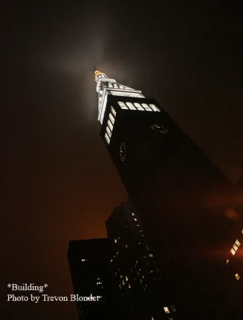 Nighttime architectural photograph by Trevon Blondet showing an upward view of a lit building against dark sky with warm amber tones.