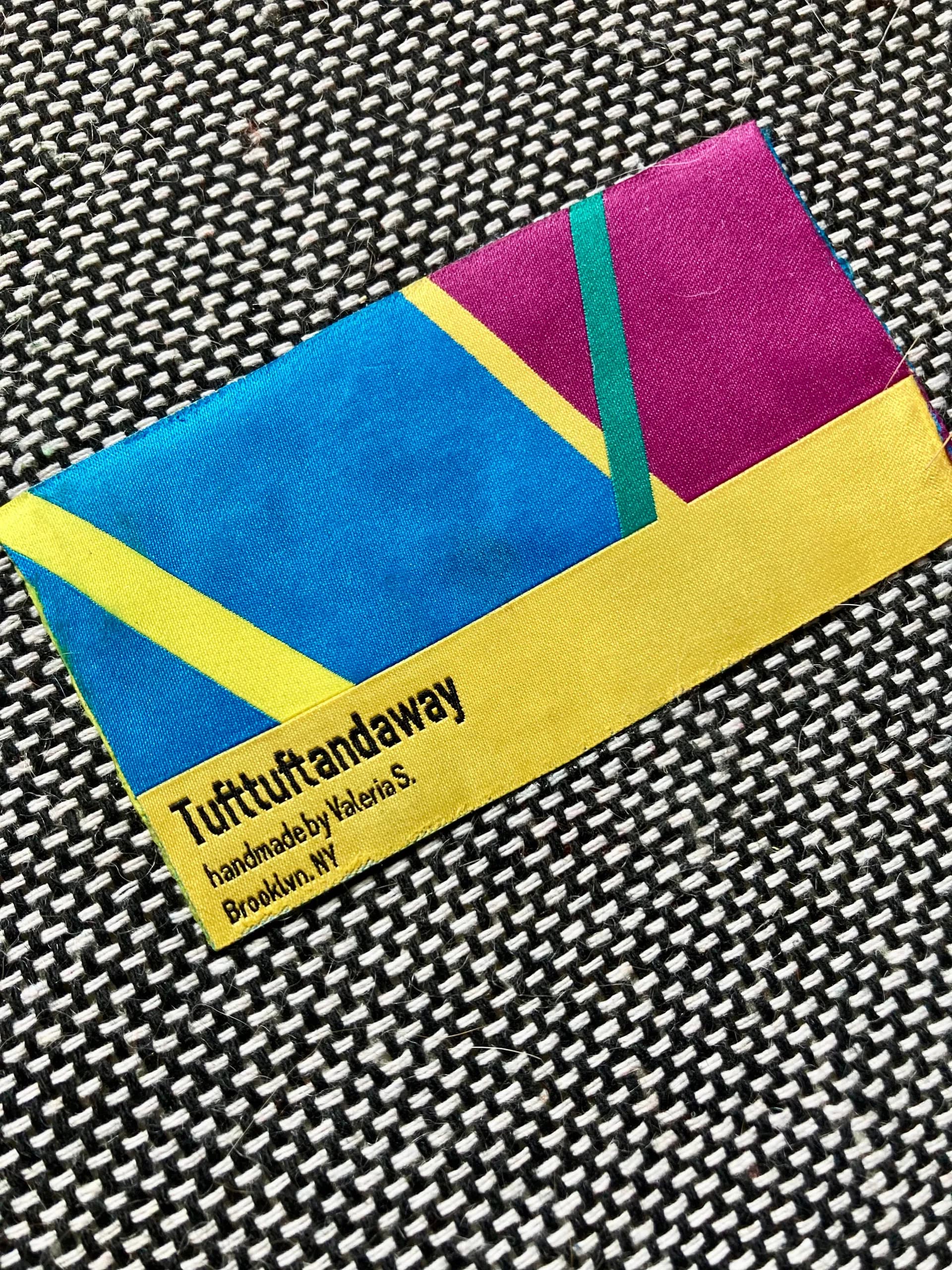 Colorful folded textile or fabric piece by Valeria Servranckx in blue, magenta, and yellow with text label, displayed on patterned background.
