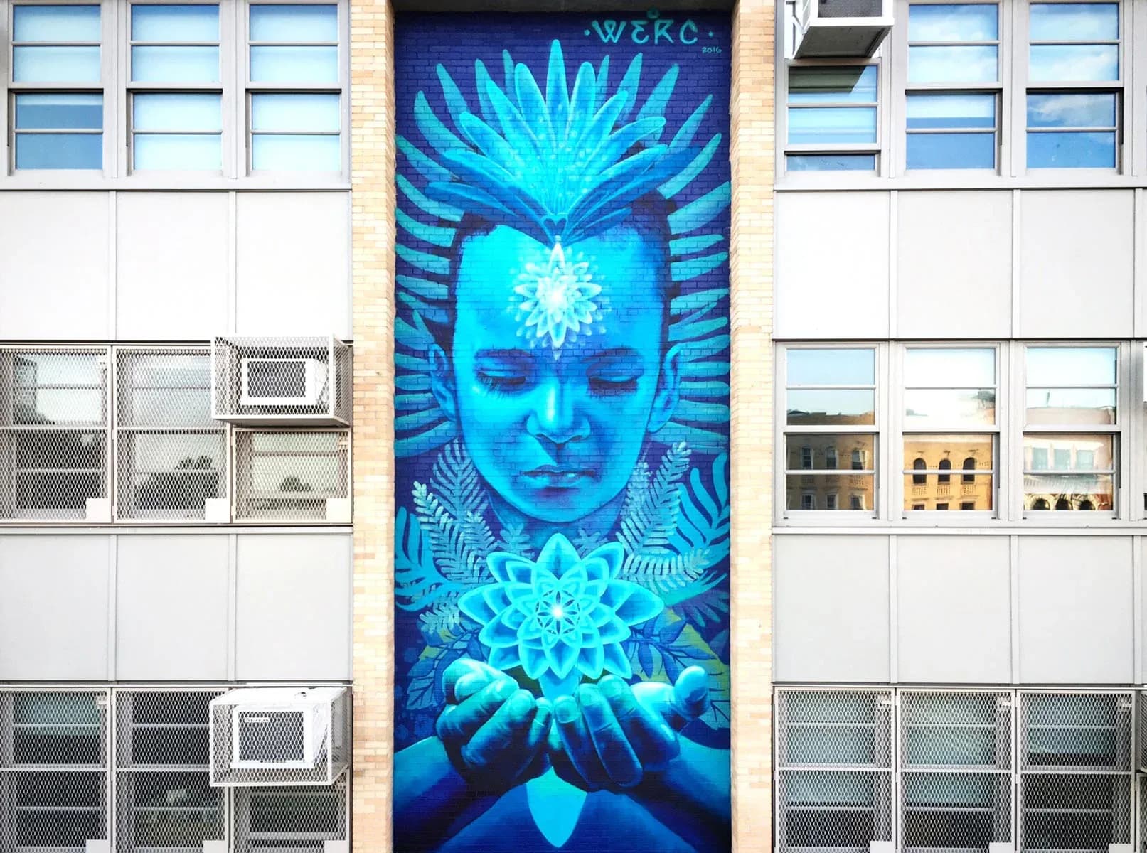 Vertical mural of a figure with feathered headdress holding a glowing mandala flower, painted in blue tones on a brick wall
