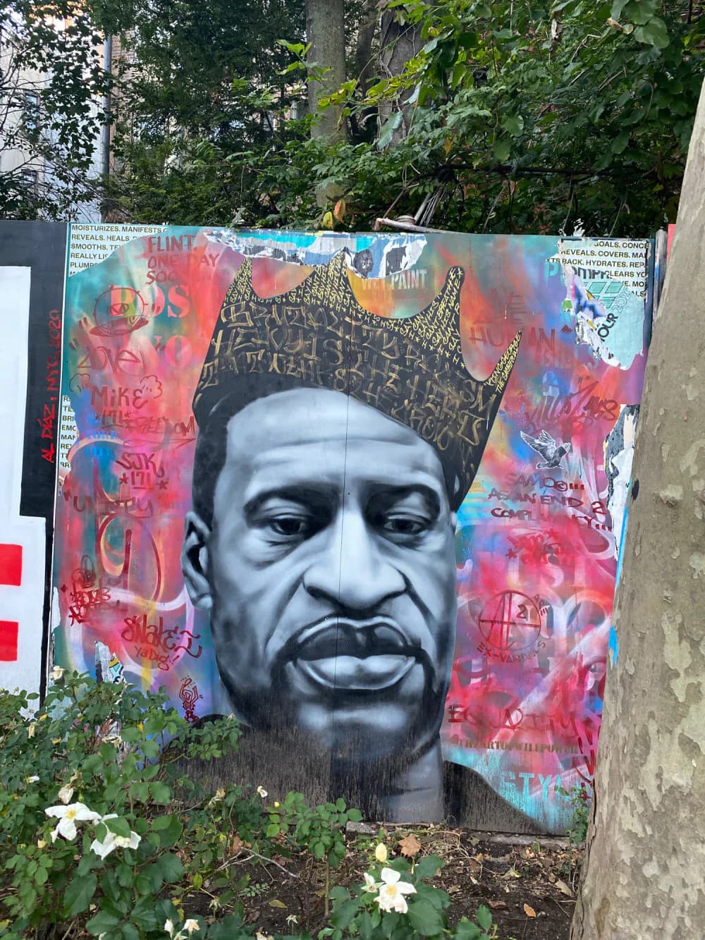 Mural portrait of a man's face in grayscale with a decorative crown, surrounded by colorful pink and blue layered paint drips and graffiti marks.
