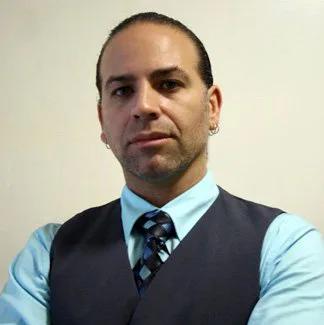 Portrait headshot of a man wearing a light blue shirt, dark vest, and patterned tie against a white background.