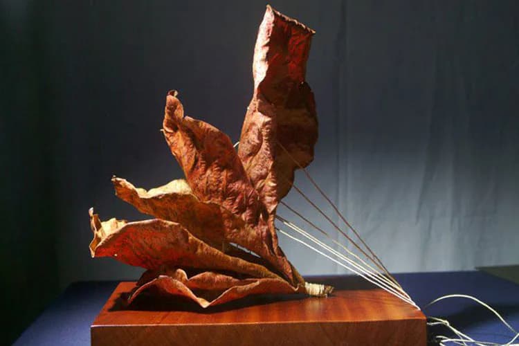 Mixed-media sculpture of a contorted human figure in copper and bronze tones mounted on a wooden base, with wire elements extending outward.