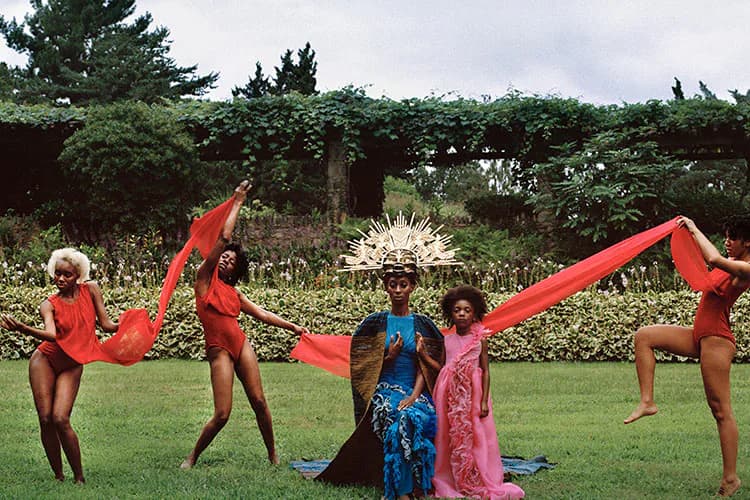 Five people in red clothing perform a dynamic outdoor installation with flowing red fabric in a grassy garden setting.