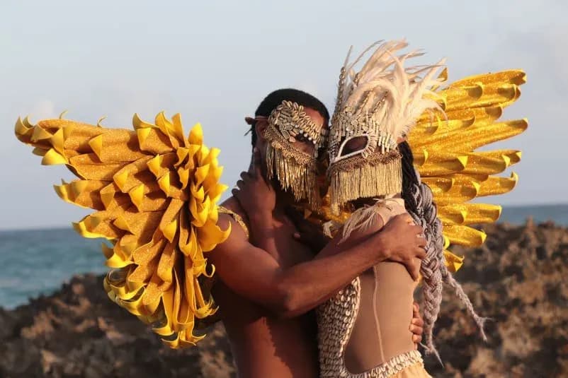 Person wearing elaborate golden wing-like structures and a textured mask poses against a rocky ocean backdrop.