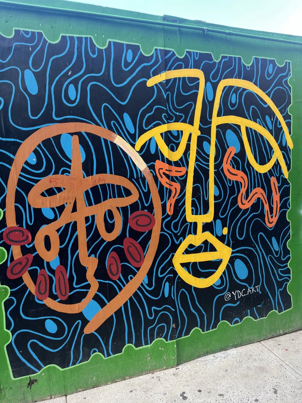 Vibrant mural by Yeline Del Carmen featuring two stylized faces in orange and yellow with expressive features against a dark blue organic patterned background on a green wall.