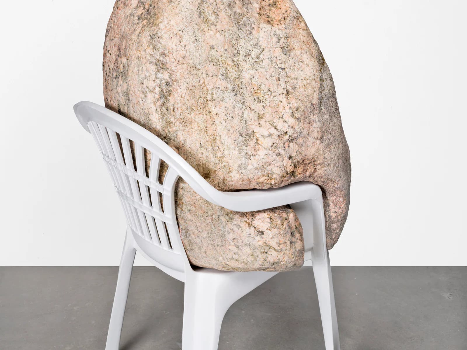 Large weathered boulder balanced on a white plastic chair, creating a surreal sculptural installation.