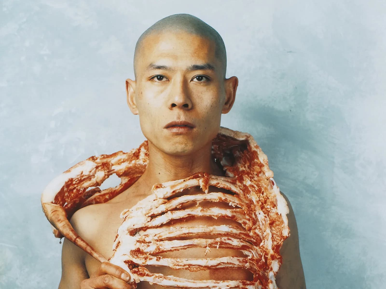Portrait of a shirtless figure wearing a textured red and white garment resembling anatomical musculature across shoulders and torso.